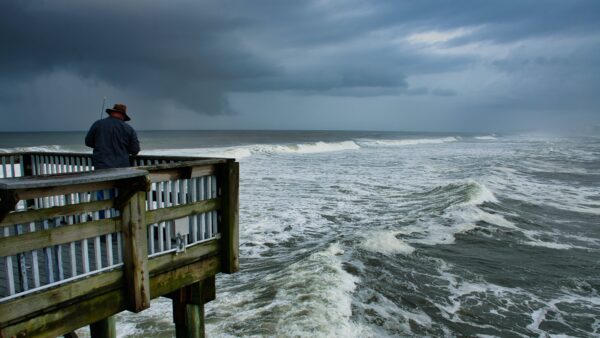 Man standing on fishing pier next to stormy skies and strong winds in the ocean.