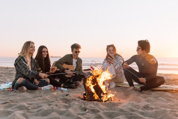 a group of friends on a beach at sunset playing music and roasting hot dogs over a campfire