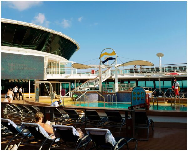 pool deck of a large cruise ship with people walking next to pool and woman sitting in lounge chair reading a book next to other empty lounge chairs