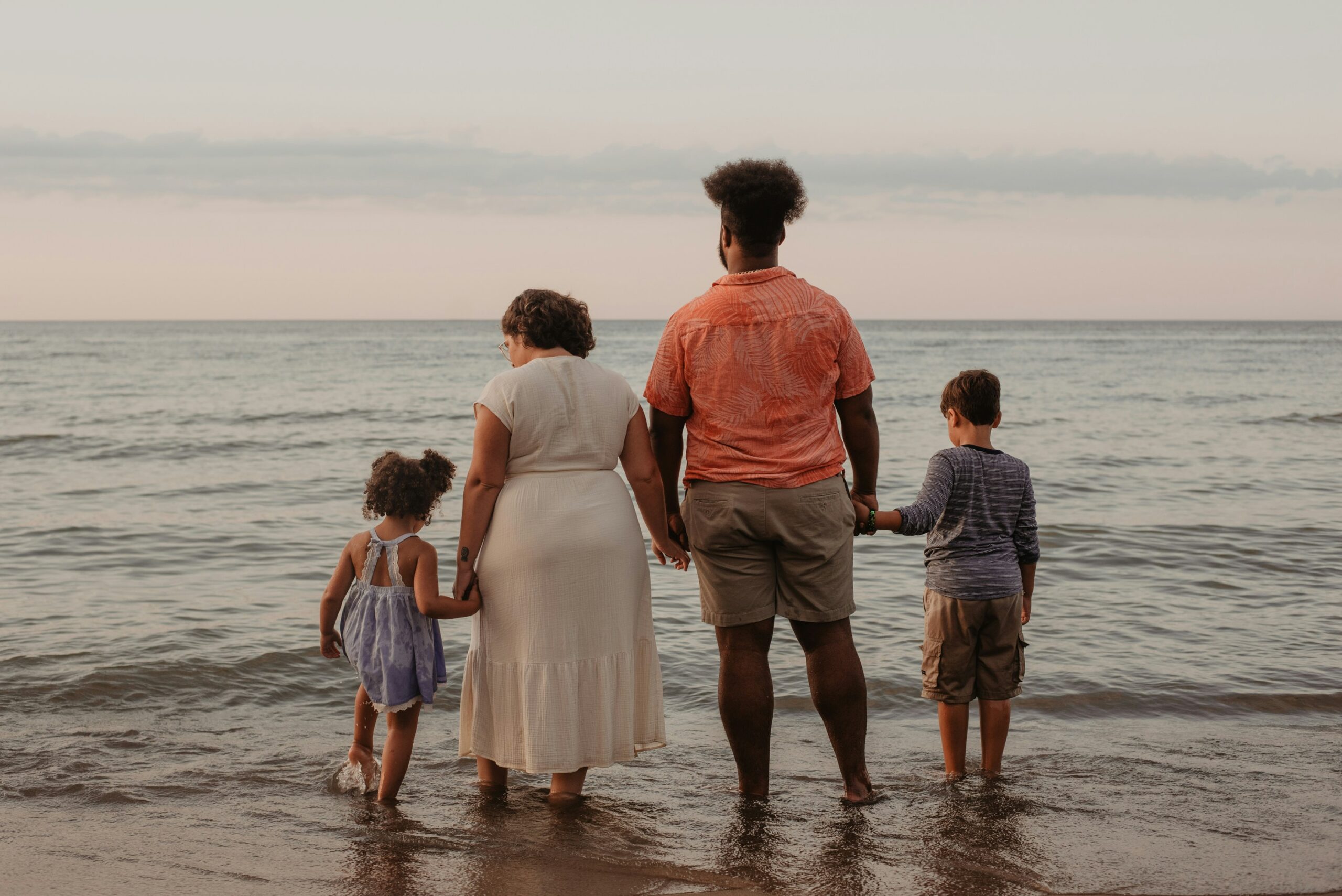 a family of four african americans with two small children all holding hands in a line facing away from the camera standing with their feet in the shallow ocean water